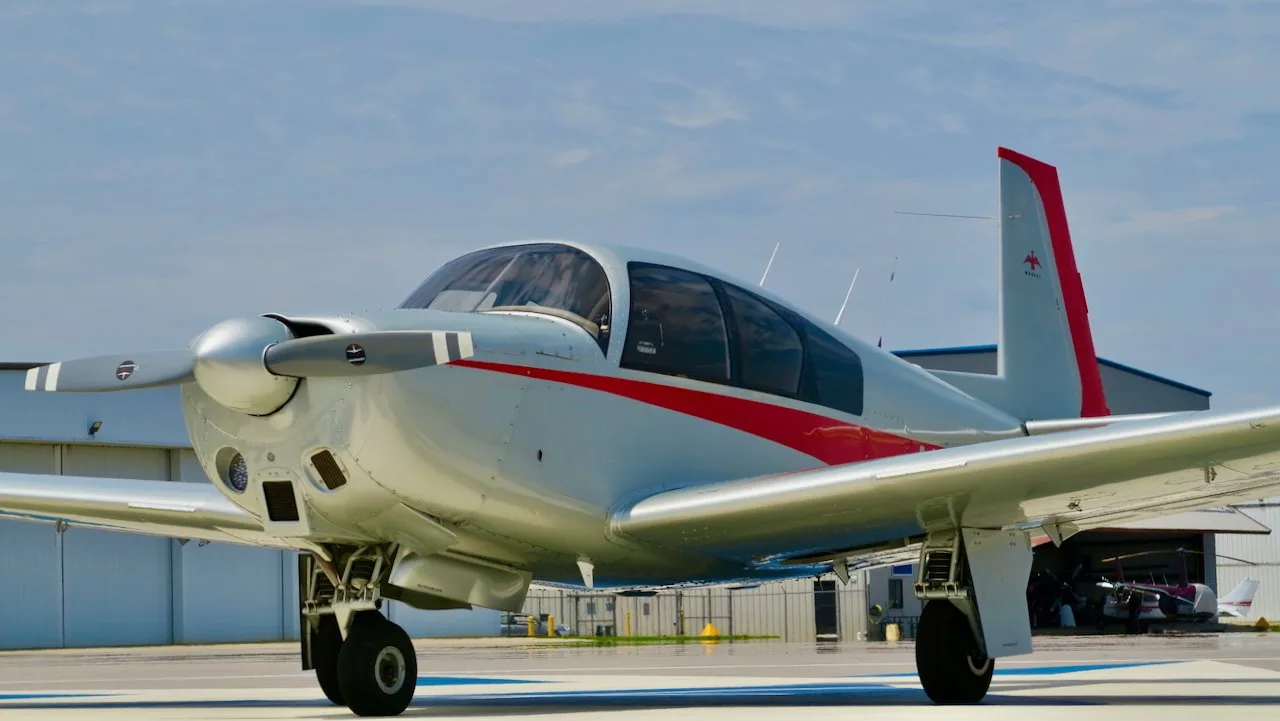 Front-right view of a silver and red Mooney aircraft parked on an airfield with hangars in the background.