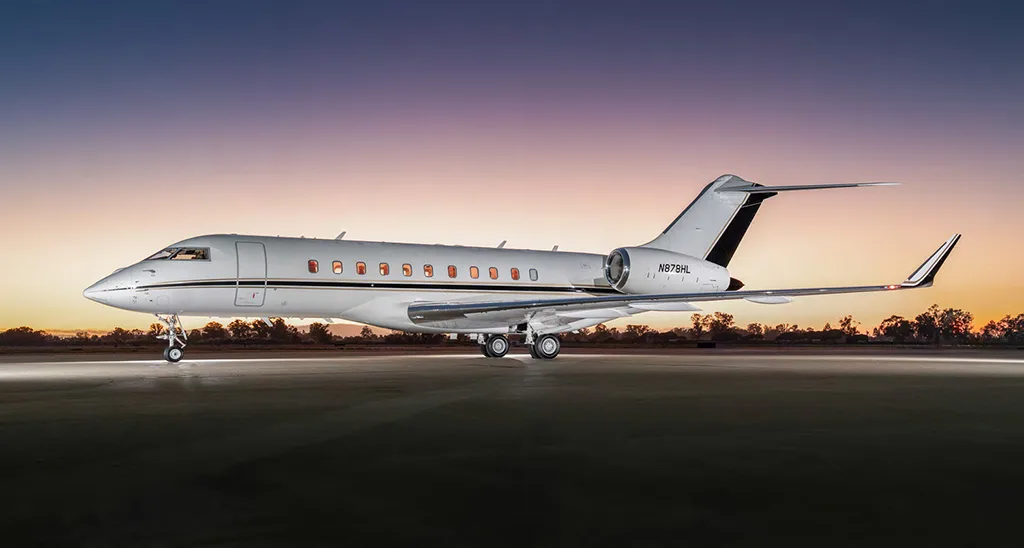 A white Bombardier Global 6000 private jet with black and gold accents on the tail, registration N878HL, parked on a runway at sunset.