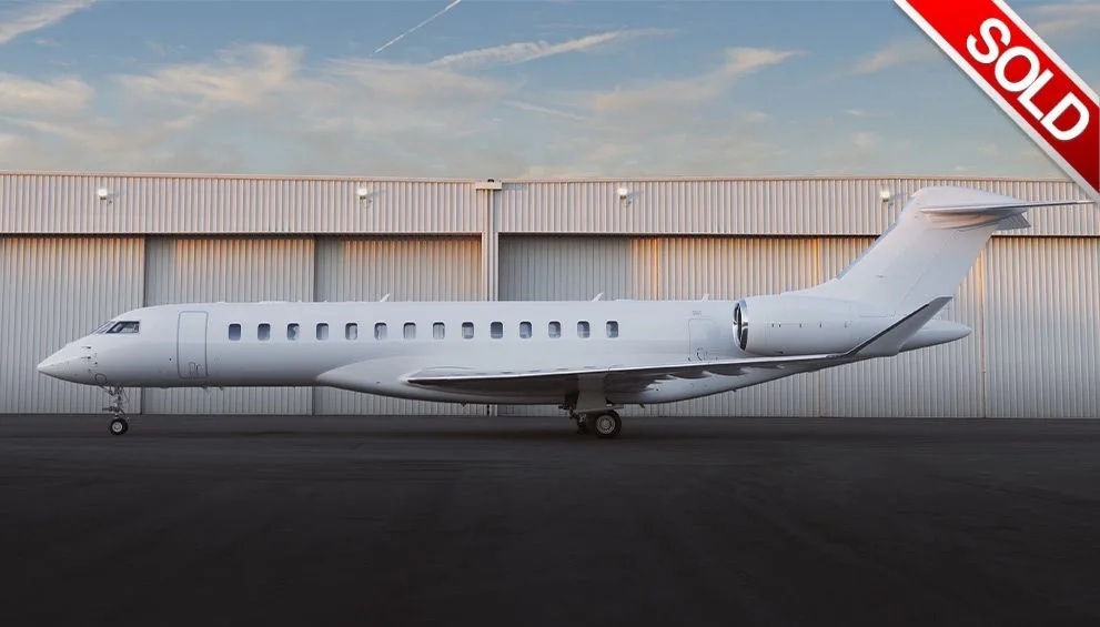 A white Bombardier Global business jet is parked on a tarmac in front of a hangar under a partly cloudy sky.