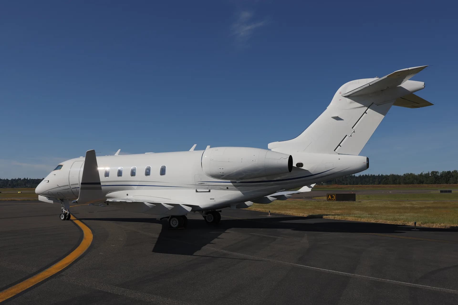 A white Bombardier Challenger business jet parked on an airport tarmac under a clear blue sky.