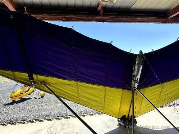 A close-up view of a purple and yellow delta-wing aircraft, with a yellow ultralight trike visible outside the hangar.