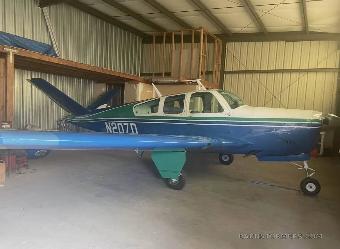 A blue and green single-engine propeller aircraft (N207D) with its wings detached, stored inside a metal hangar.
