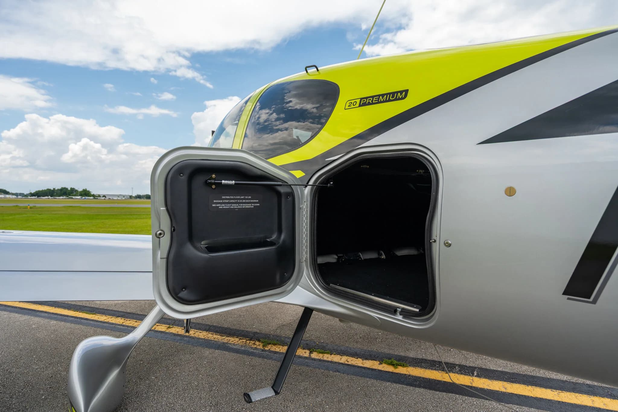 Close-up of the open baggage compartment on the left side of a lime green and silver Cirrus SR20 aircraft, with "20 PREMIUM" visible on the fuselage.