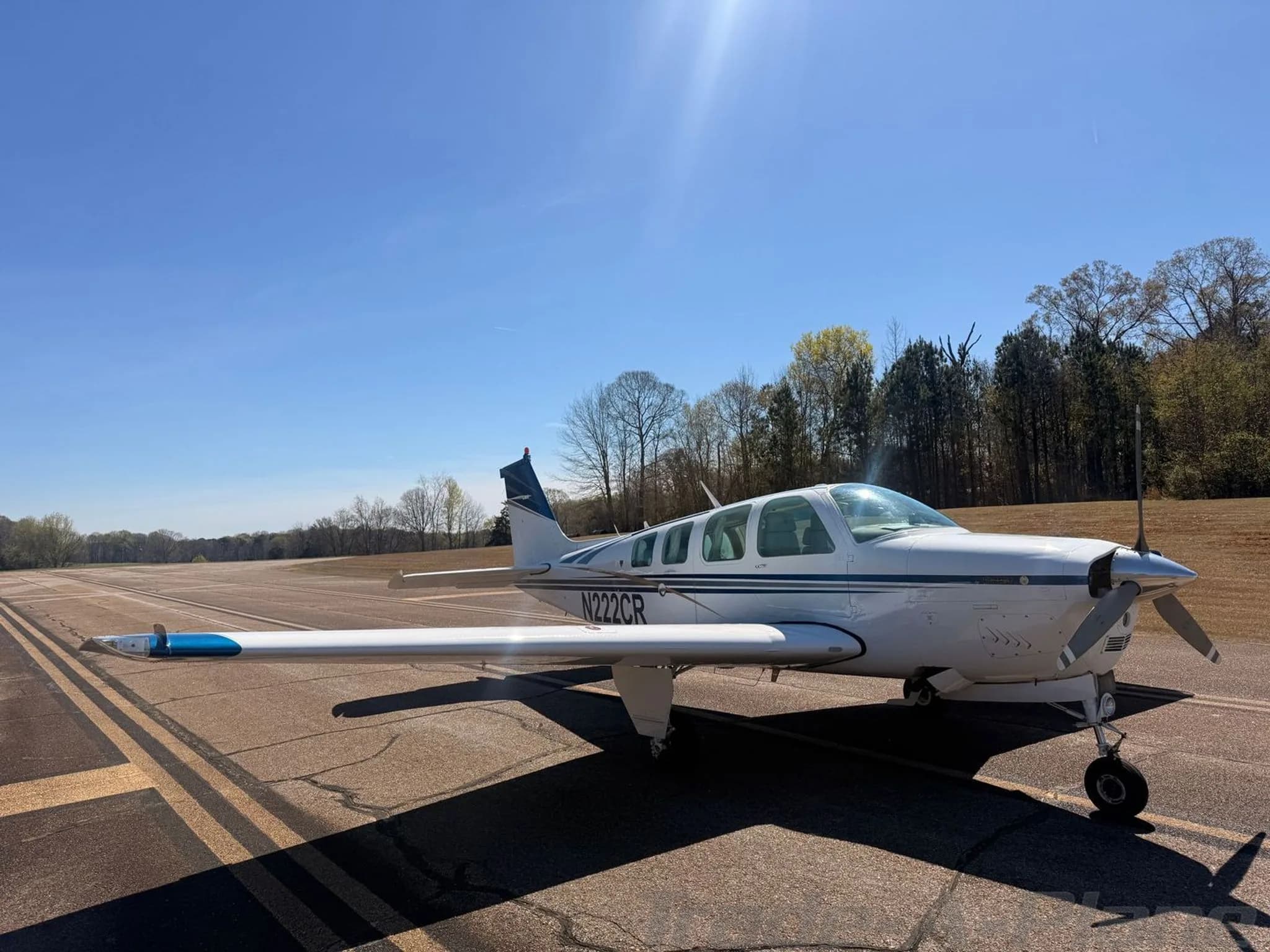 A white Beechcraft Bonanza N222CR with blue and gold stripes parked on an asphalt runway, viewed from the front-right.