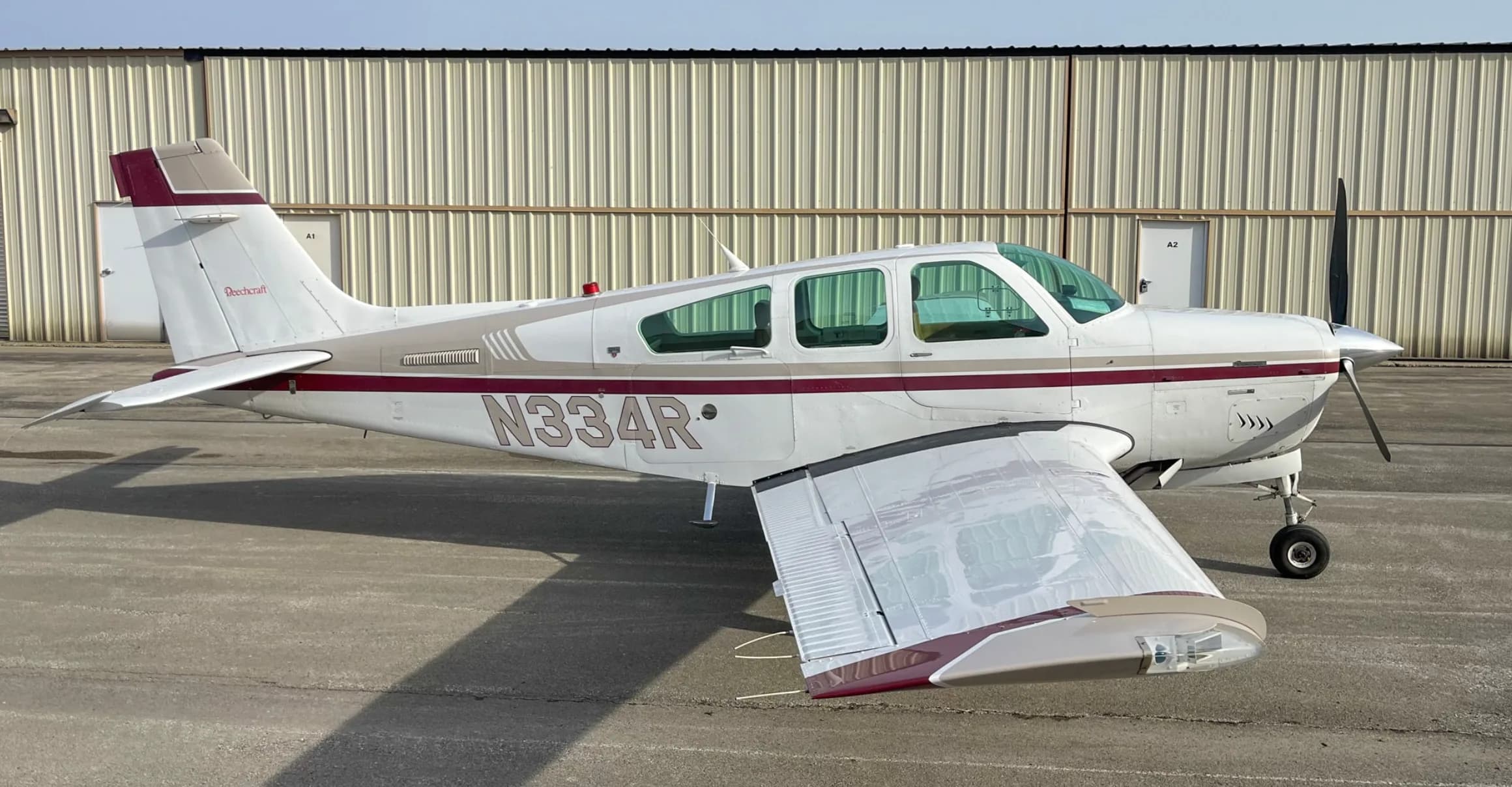 A side view of a white and maroon Beechcraft Bonanza aircraft, tail number N334R, parked in front of a hangar.