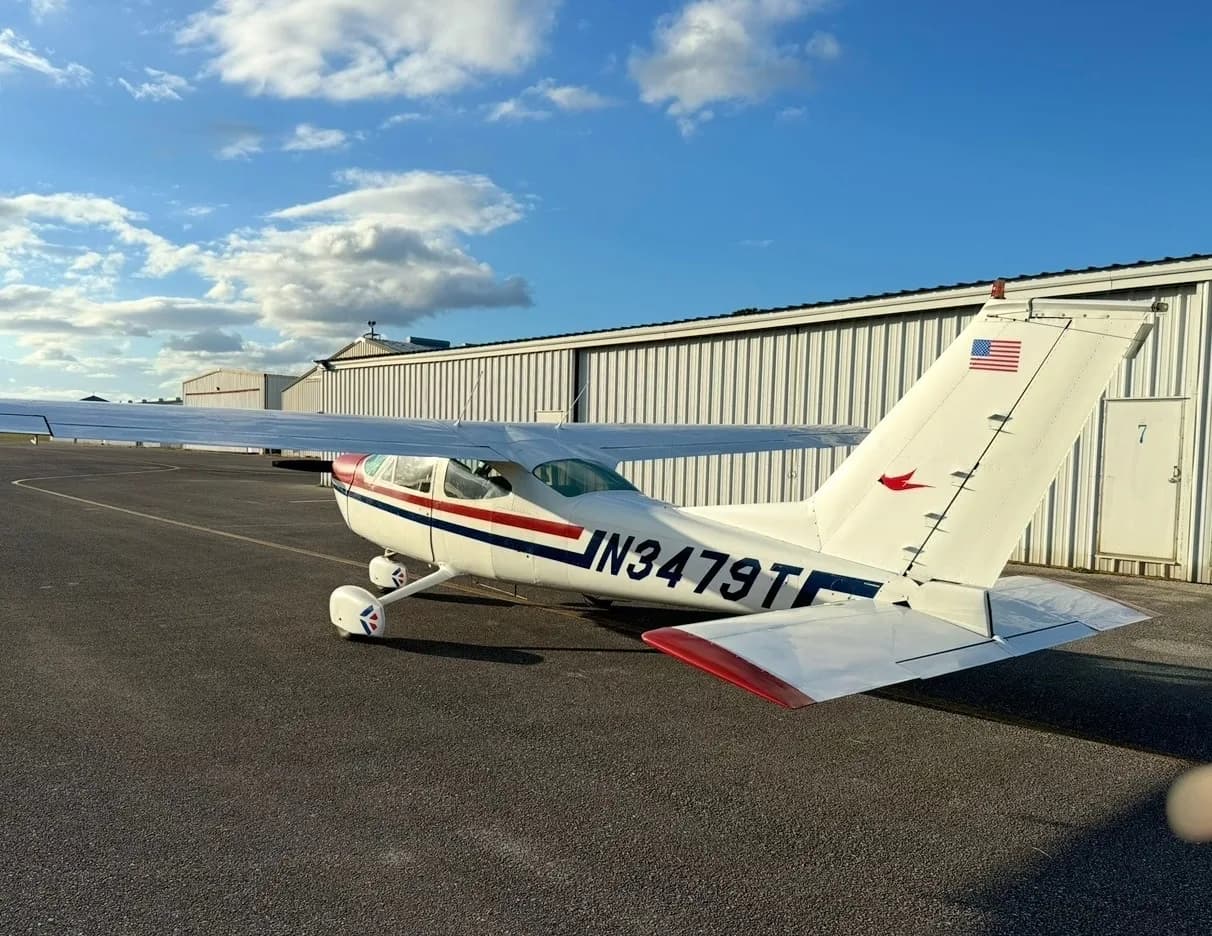 A white Cessna 172 with red and blue stripes and a US flag on its tail is parked on an asphalt ramp next to a hangar.