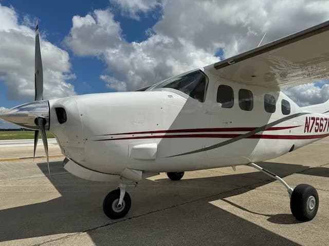 A front-side view of a white Cessna 206 with red and grey stripes, showing the propeller and N-number N7567K, parked on an airfield.
