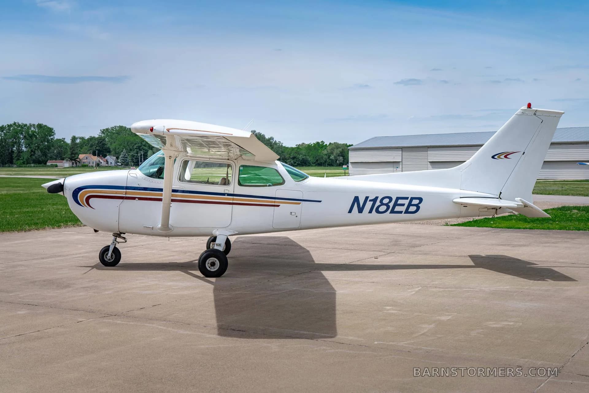 A white Cessna 172 (N18EB) with blue and gold stripes is parked on a tarmac, viewed from the right side.