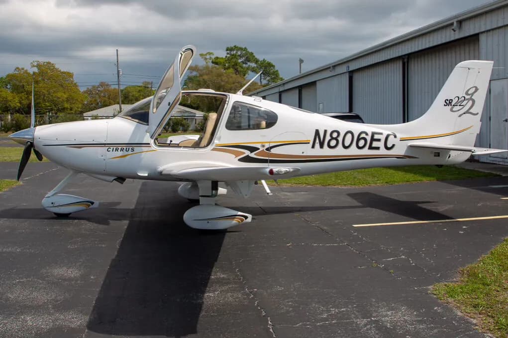 A white Cirrus SR22 aircraft, registration N806EC, with its left gull-wing door open, parked on an asphalt tarmac.