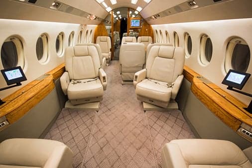 A view down the aisle of a private jet cabin with cream leather seating and individual entertainment screens.