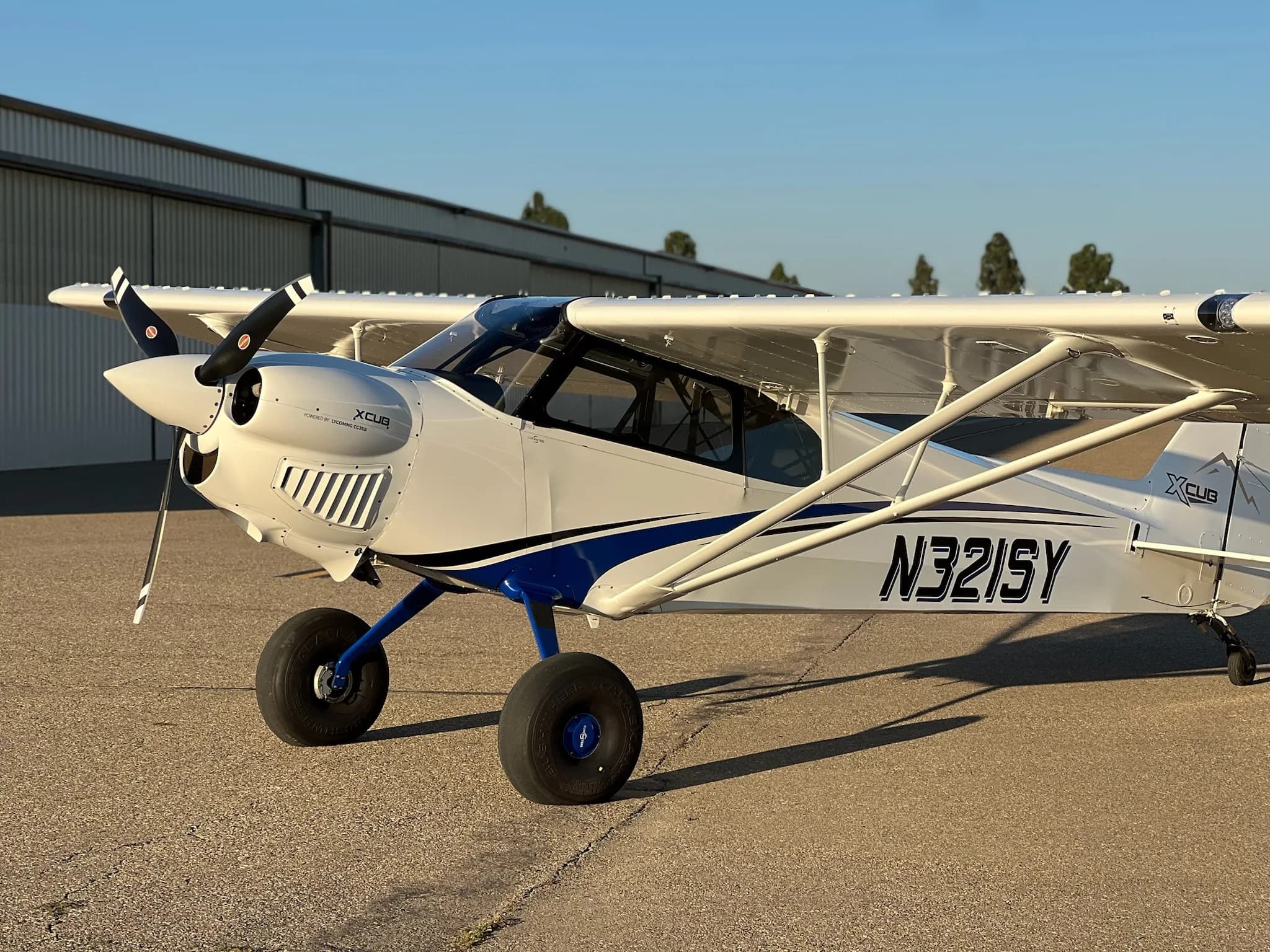 A front-right view of a white X-Cub aircraft with blue stripes and registration N321SY, parked on an airfield.