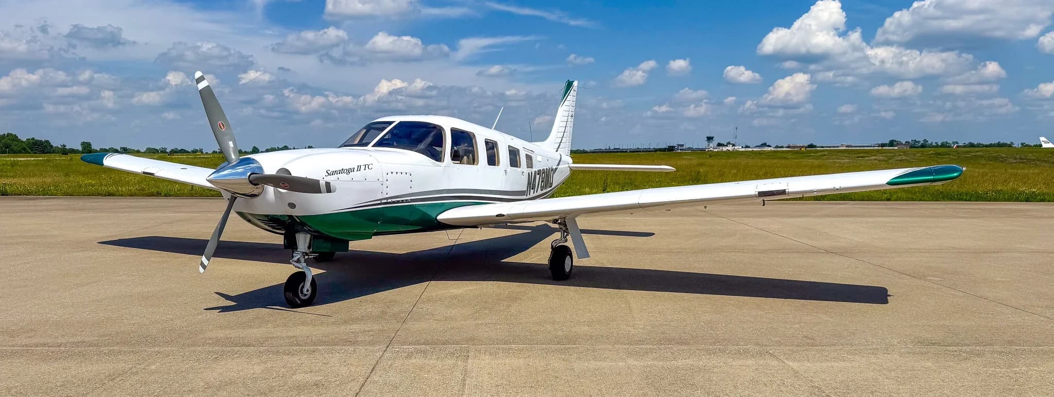 A white and green Piper Saratoga II TC aircraft (N478MD) parked on an airfield with distant buildings and trees.