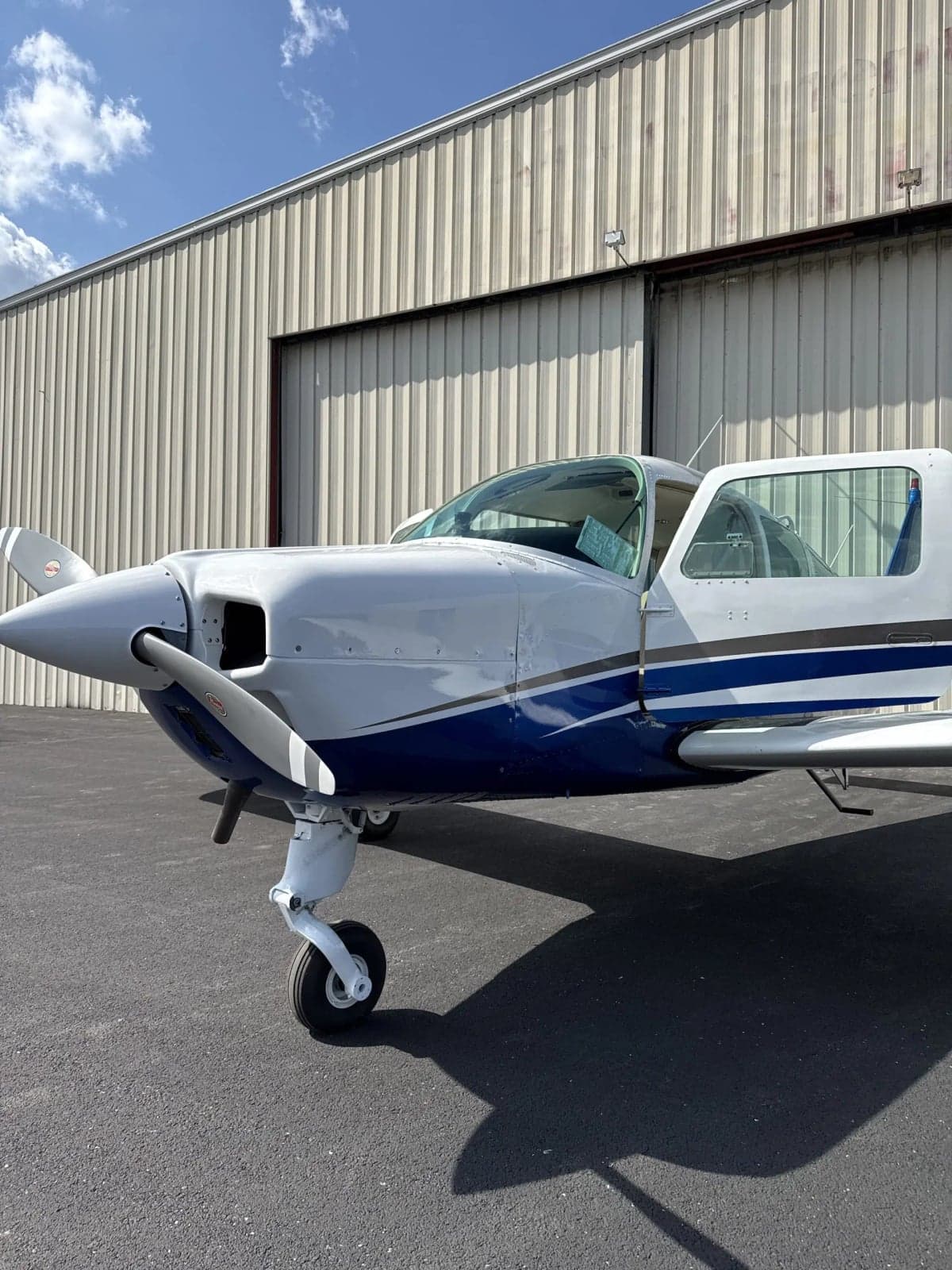 A white and blue single-engine aircraft with its door open, parked on the tarmac in front of a hangar.