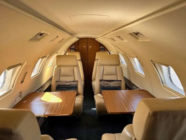 The aircraft's cabin interior features beige leather seats, two facing pairs with wooden tables in between, and overhead lighting.