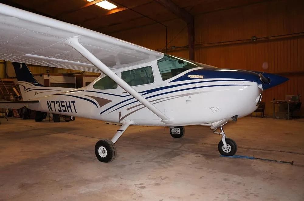 A white and blue Cessna 182 Skylane N735HT is parked in a hangar, viewed from the front-right.