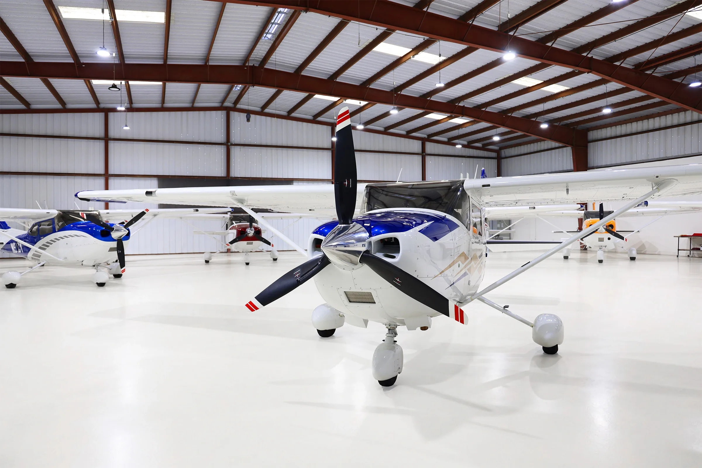 A front view of a white and blue Cessna Skylane with gold accents, parked in a hangar with other aircraft.