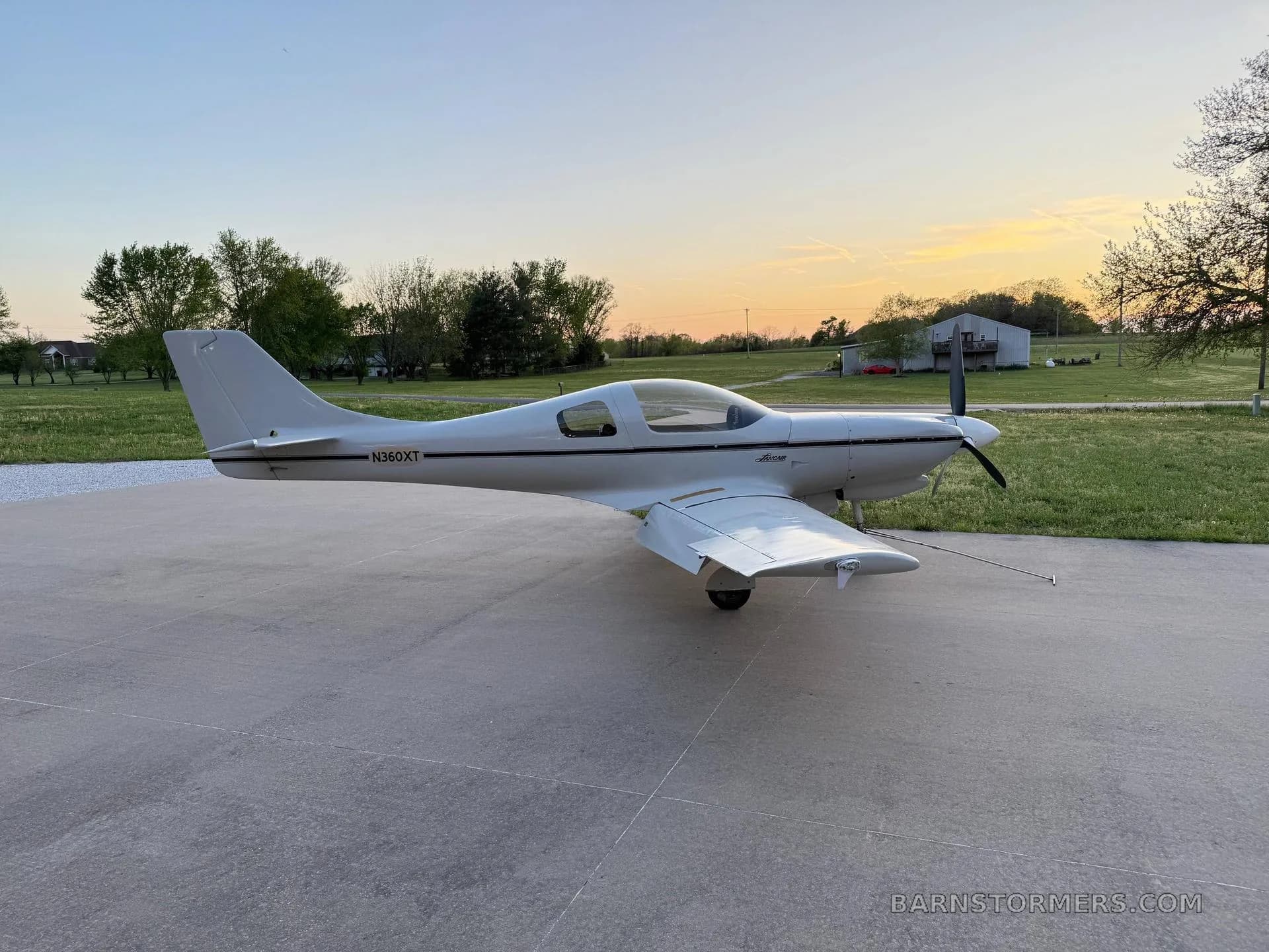 A white Lancair Legacy aircraft (N360XT) with a black stripe is parked on a concrete pad at sunset, viewed from the side.