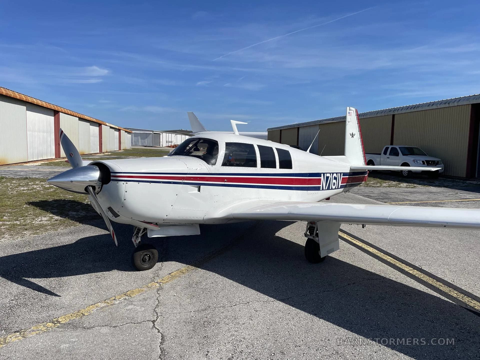 A white single-engine propeller aircraft with red and blue stripes on the fuselage and tail, parked on an asphalt ramp with hangars in the background.