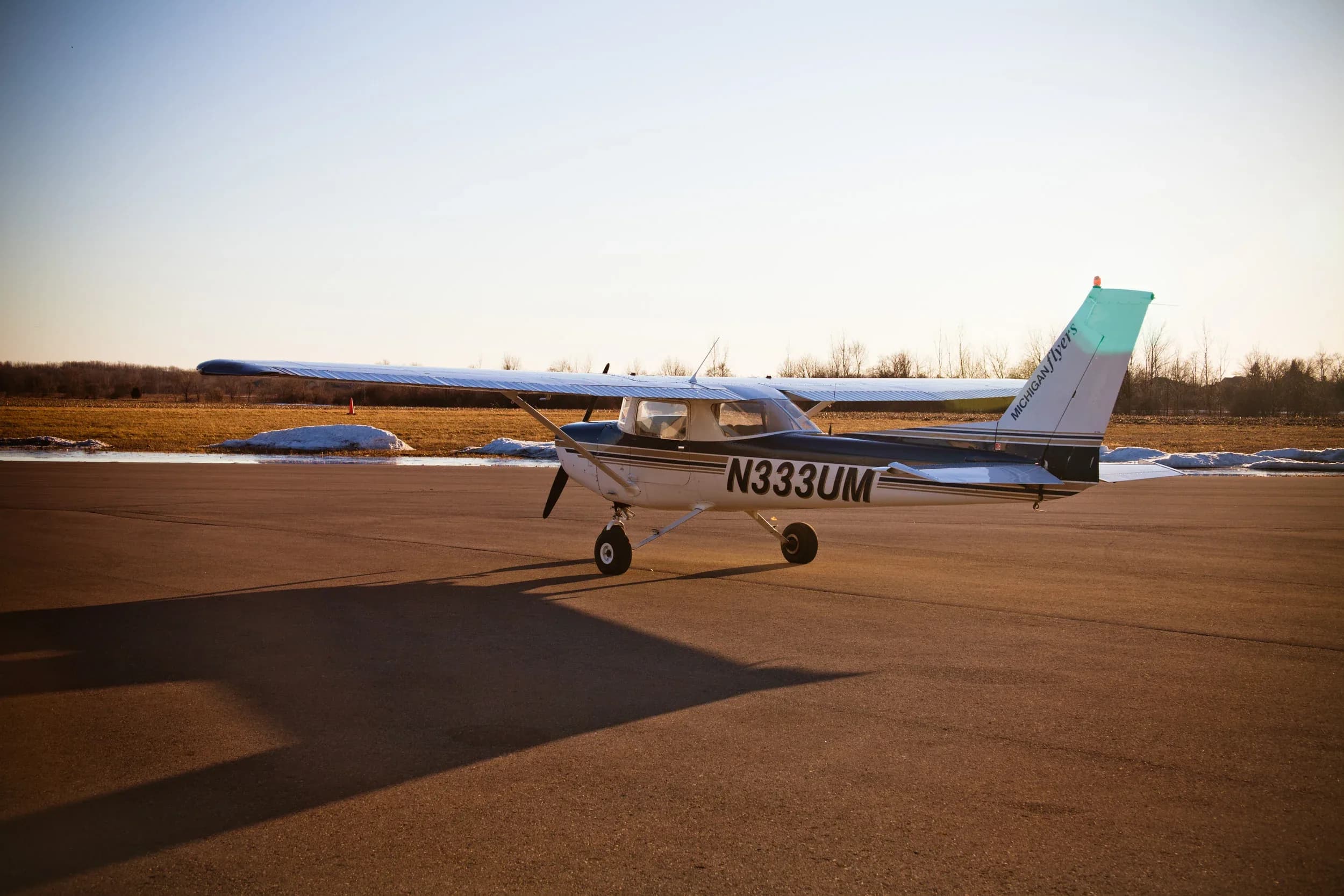 A white and dark blue Cessna-like aircraft with registration N333UM is parked on a tarmac at sunset, casting a long shadow.