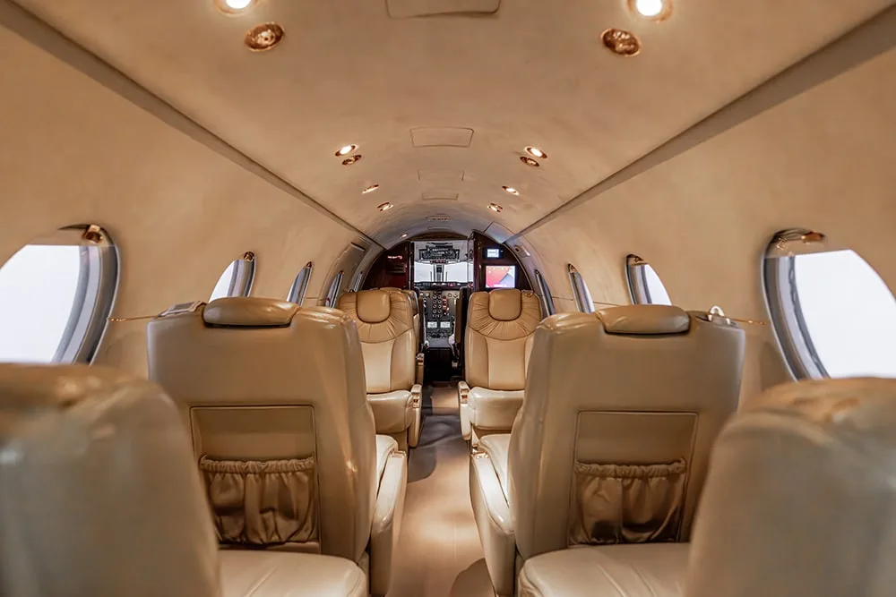View down the aisle of a private jet cabin with light-colored leather seats and the cockpit visible in the distance.