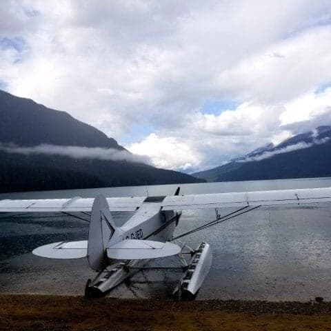 A white and blue Super Cub floatplane on the shore of a lake with mountains in the background.