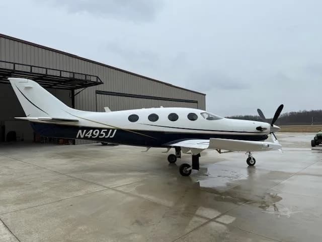 A white and dark blue Daher TBM aircraft, N495JJ, is parked on a wet tarmac in front of a hangar under an overcast sky.