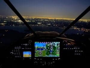 The cockpit view of an aircraft at night, with city lights visible in the distance.
