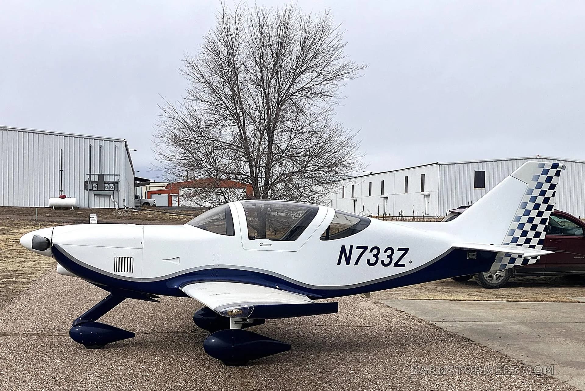 A white and blue single-engine aircraft with registration N733Z parked on an airfield with buildings and a bare tree in the background.