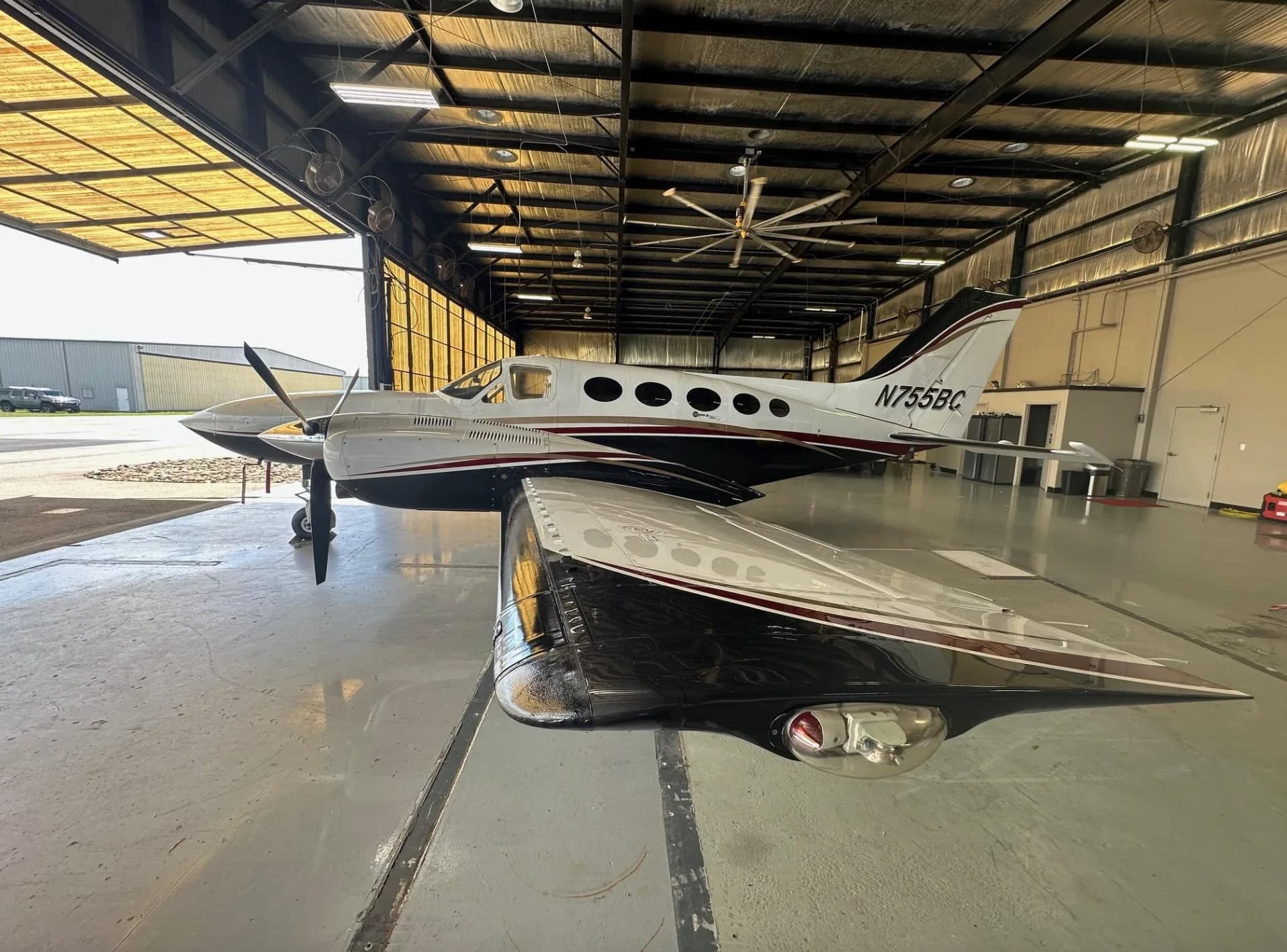 Rear-side view of a white, black, and red twin-engine turboprop aircraft, N755BC, with its wing in the foreground, inside a hangar.