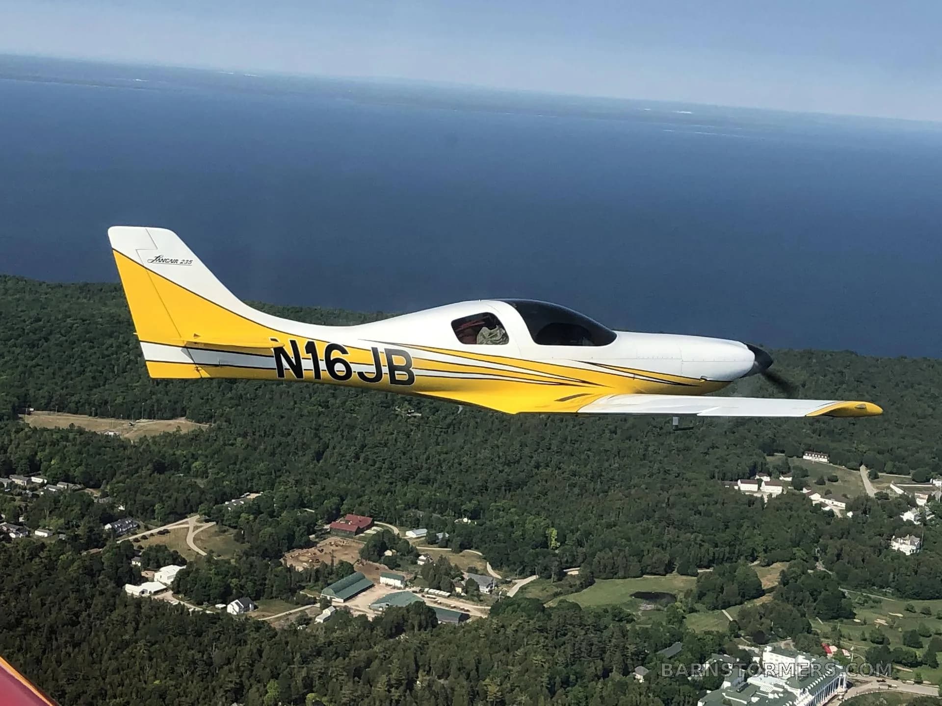 A white and yellow single-engine aircraft with tail number N16JB, flying over a forested coastline with a large body of water.