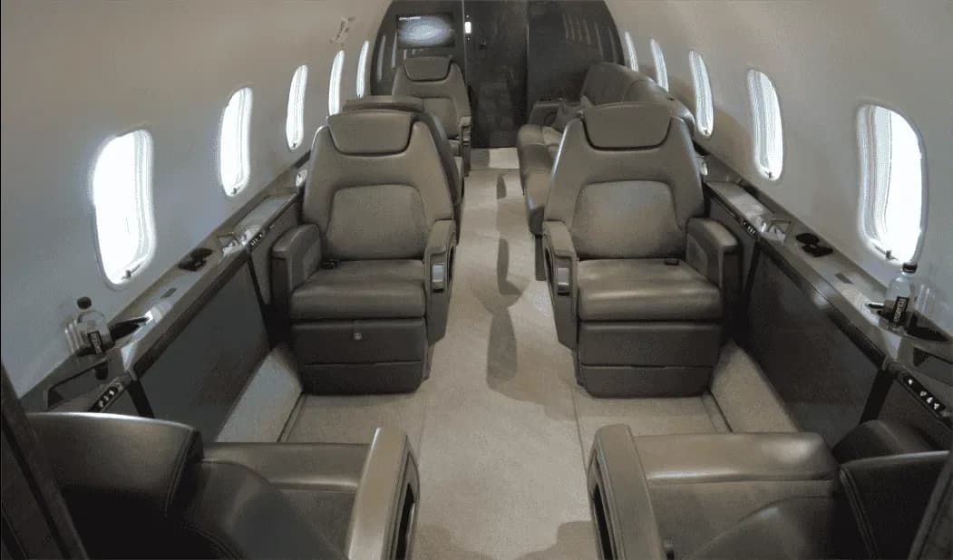 The spacious interior of a business jet cabin with multiple grey leather seats arranged in a club configuration.