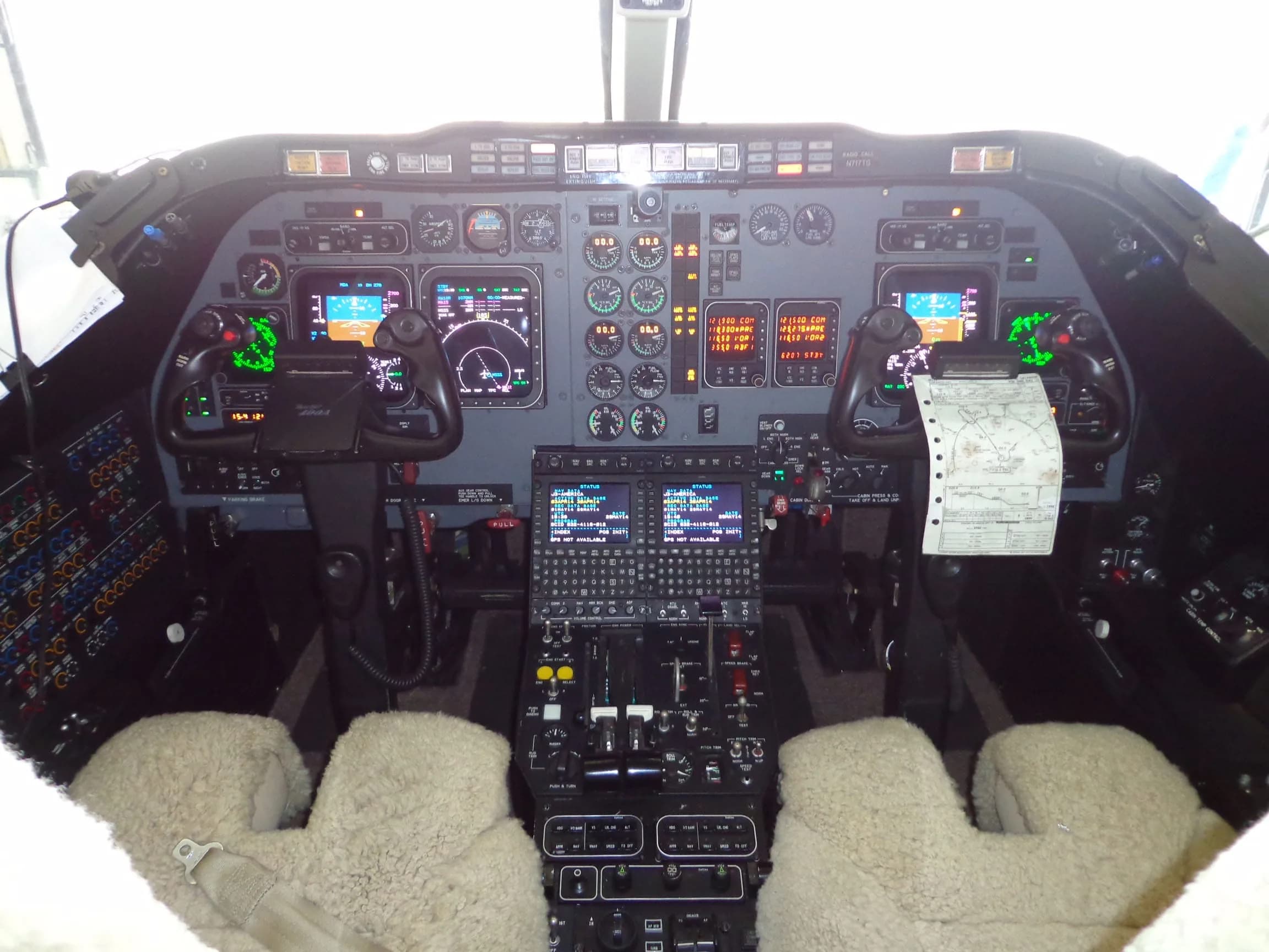 The cockpit of a private jet, featuring multiple screens, gauges, and controls with sheepskin seat covers.