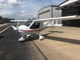 A white and red light sport aircraft is parked on the tarmac in front of a hangar.