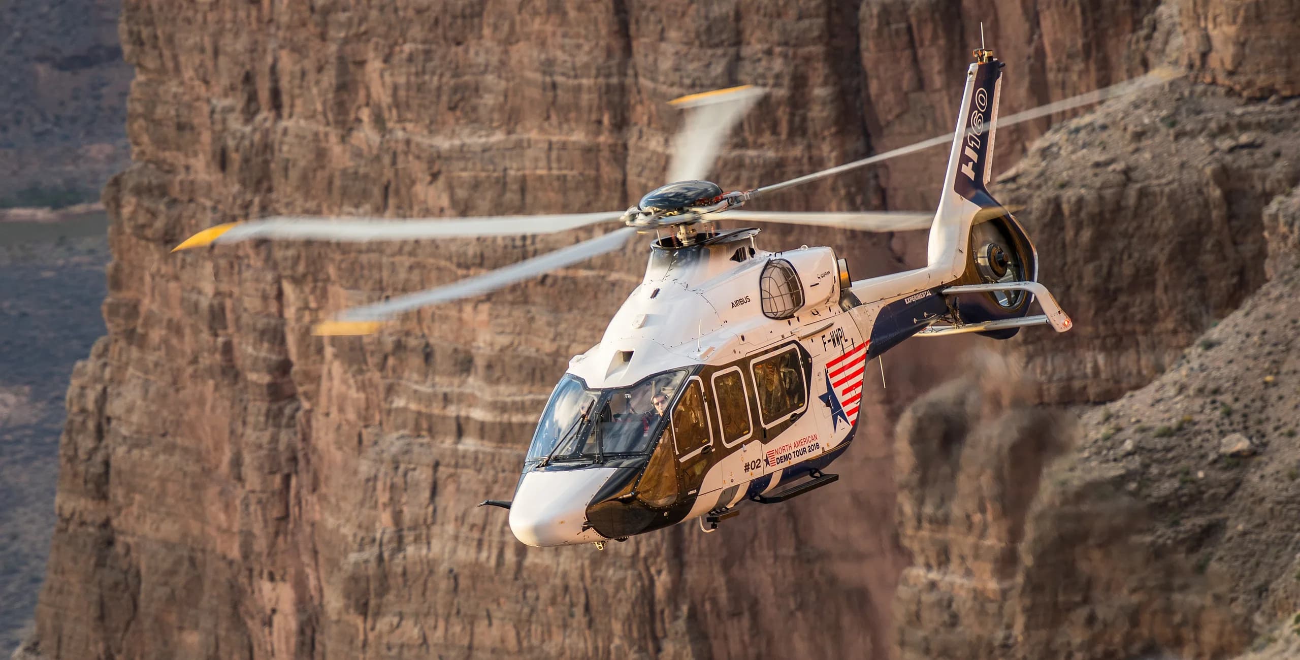 An Airbus H160 helicopter flying near a canyon wall.