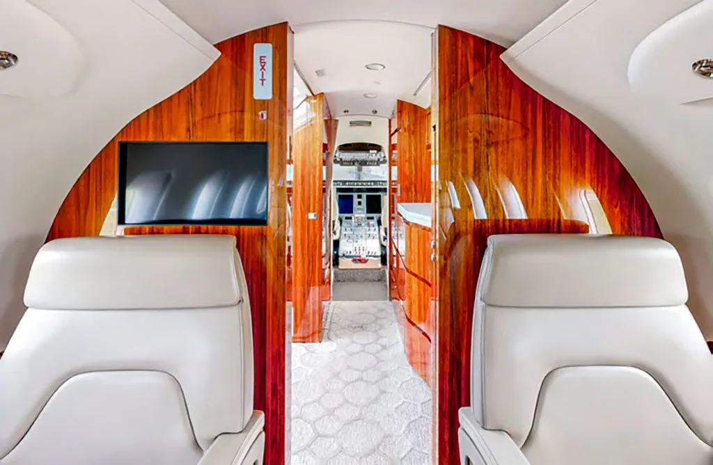 A view down the private jet cabin aisle, featuring wood paneling and a partial view of the cockpit.