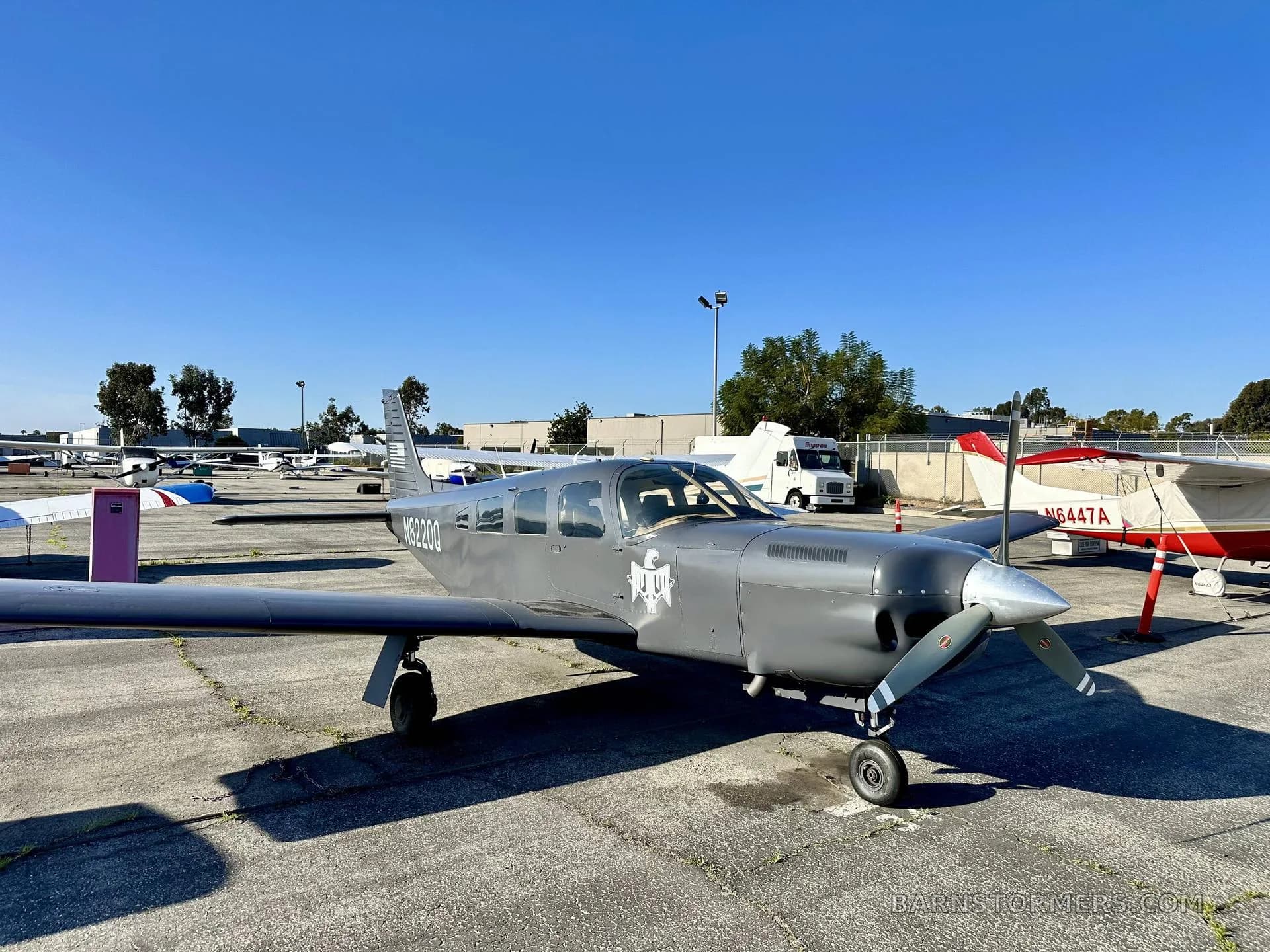 A gray single-engine propeller aircraft, N8220Q, with a white emblem on its nose, parked on a concrete tarmac under a clear blue sky.