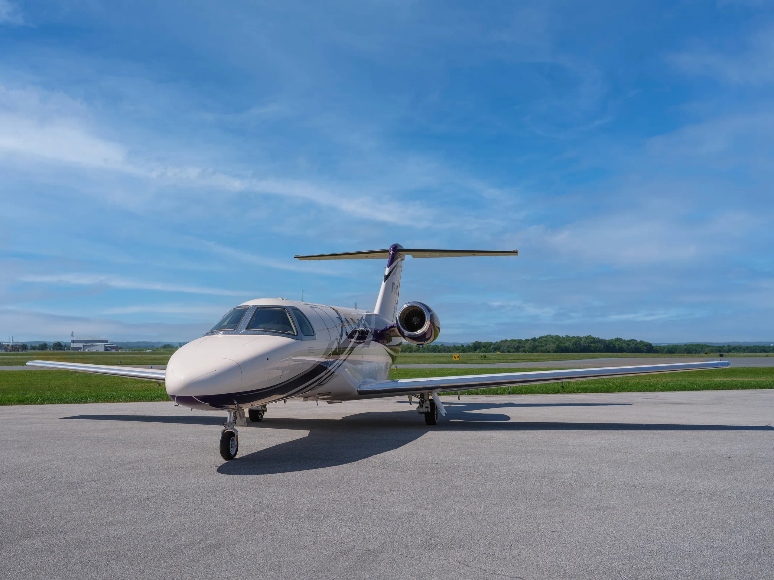 A white and purple Cessna Citation CJ4 business jet is parked on an asphalt tarmac under a blue sky with scattered clouds.