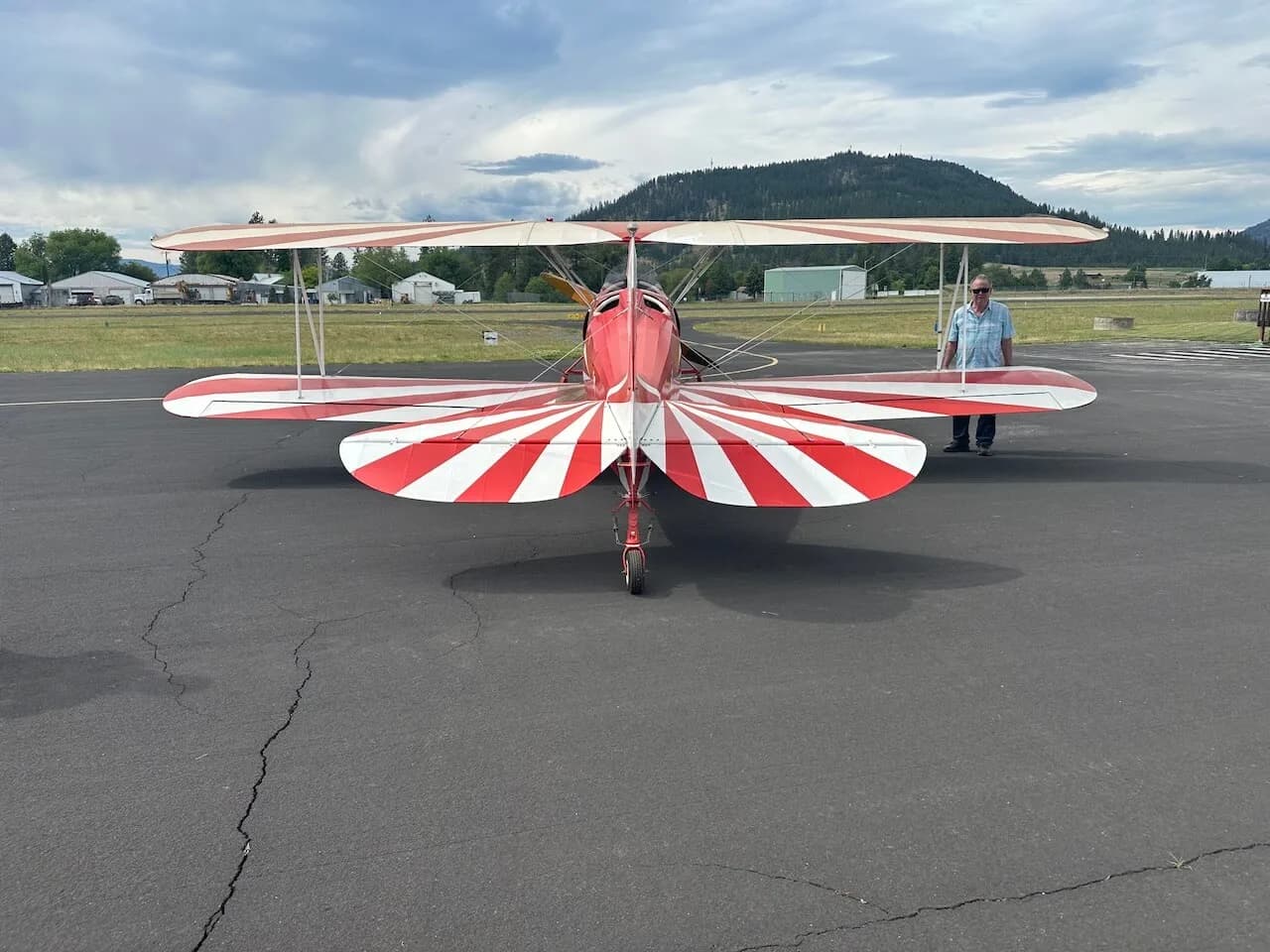 A red and white biplane with striped wings and tail, viewed from the rear, with a person standing nearby on an airfield.