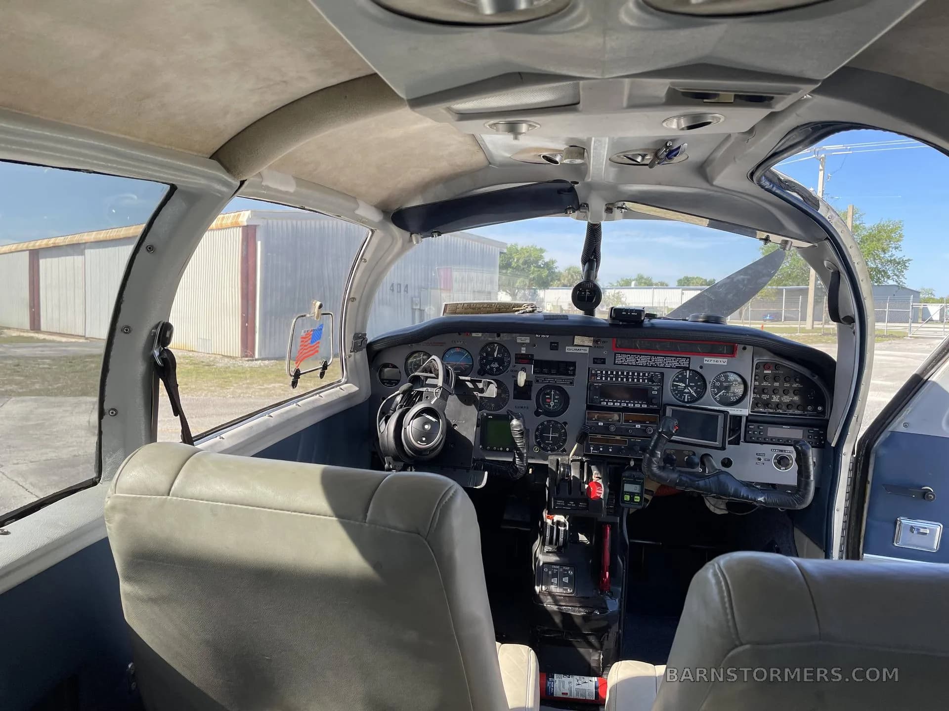 Wide view of the aircraft's cockpit, showing the instrument panel, controls, and two front seats, with hangars visible through the windshield.
