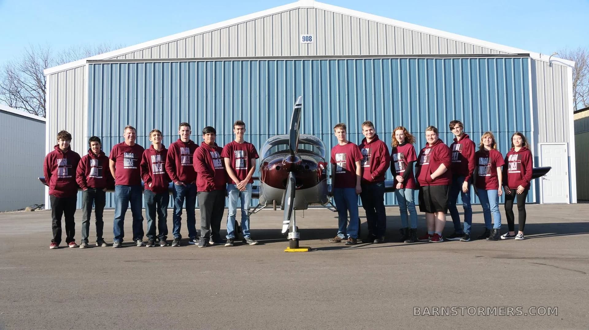 A group of students and adults in matching maroon shirts standing in front of a burgundy and silver single-engine aircraft outside a hangar.