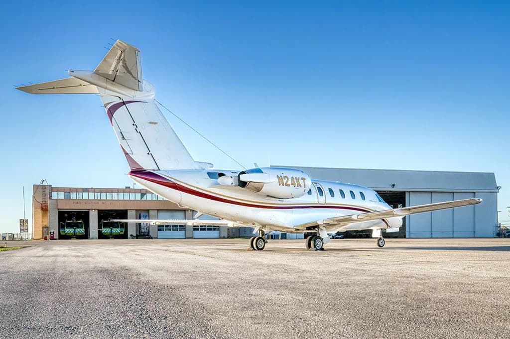 A white business jet with red and gold stripes (N24KT) is parked on a tarmac in front of a hangar.