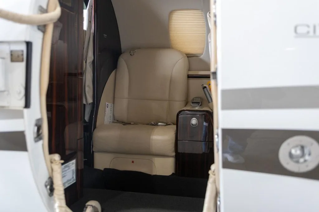 The open cabin door of a business jet revealing a single beige leather seat and dark wood paneling.