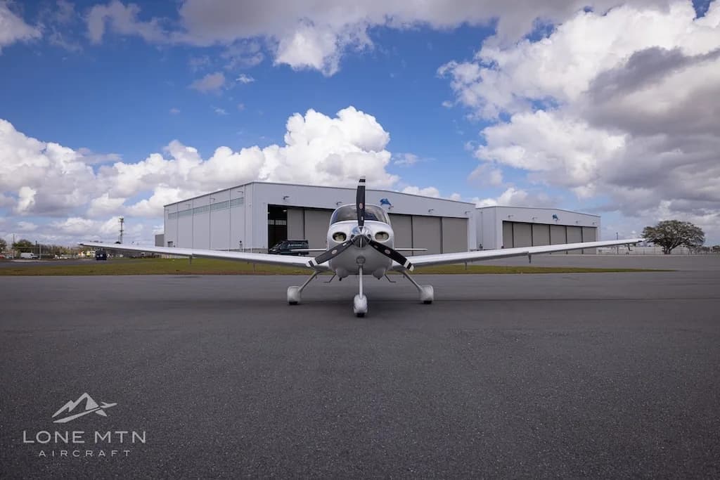 A head-on view of the white Cirrus SR20 aircraft (N166PG) parked on an airport tarmac in front of hangars.