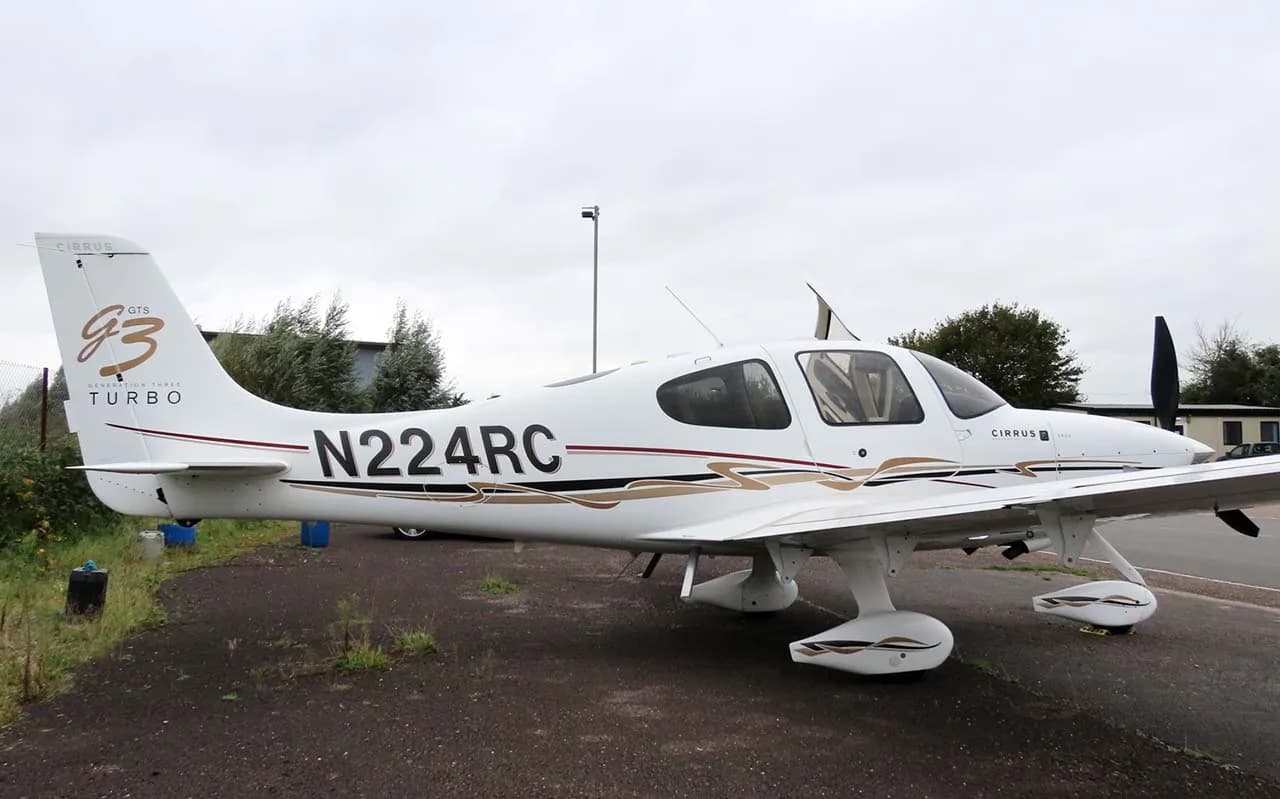 A white Cirrus SR22T (N224RC) with brown and black stripes parked on an asphalt surface next to a grassy area.