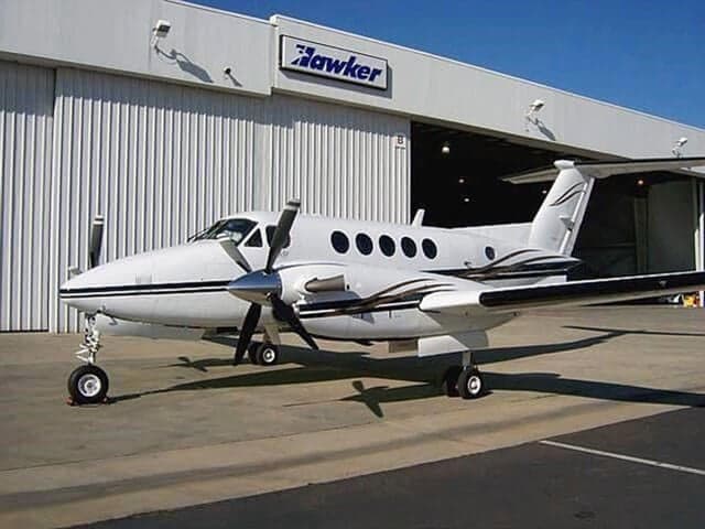 A white Beechcraft King Air with brown and black stripes is parked outside a Hawker hangar.