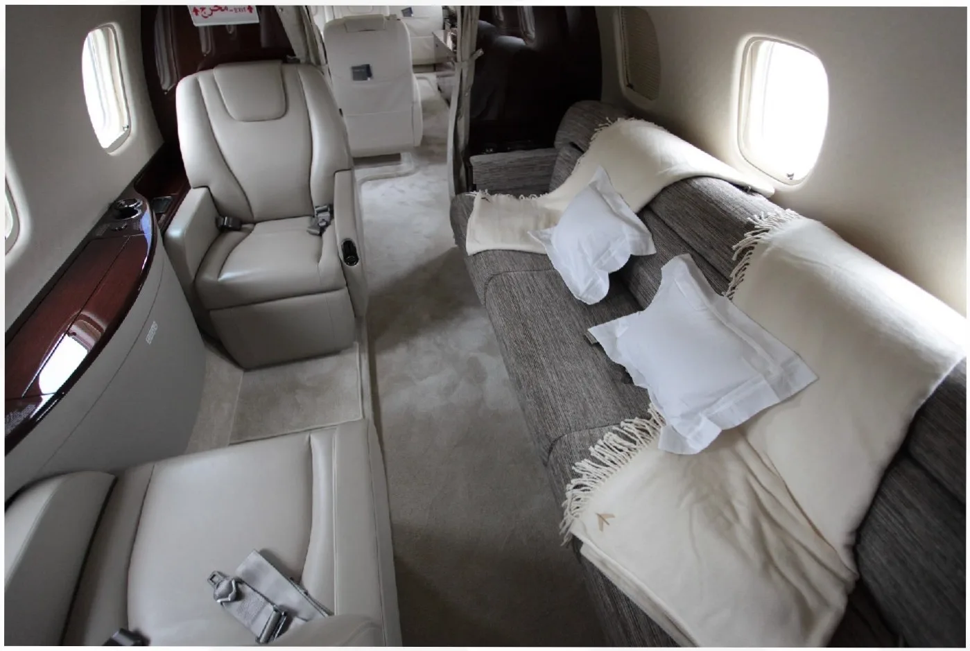 The luxurious interior of a private jet cabin features a long couch with blankets and pillows, facing individual cream-colored seats.