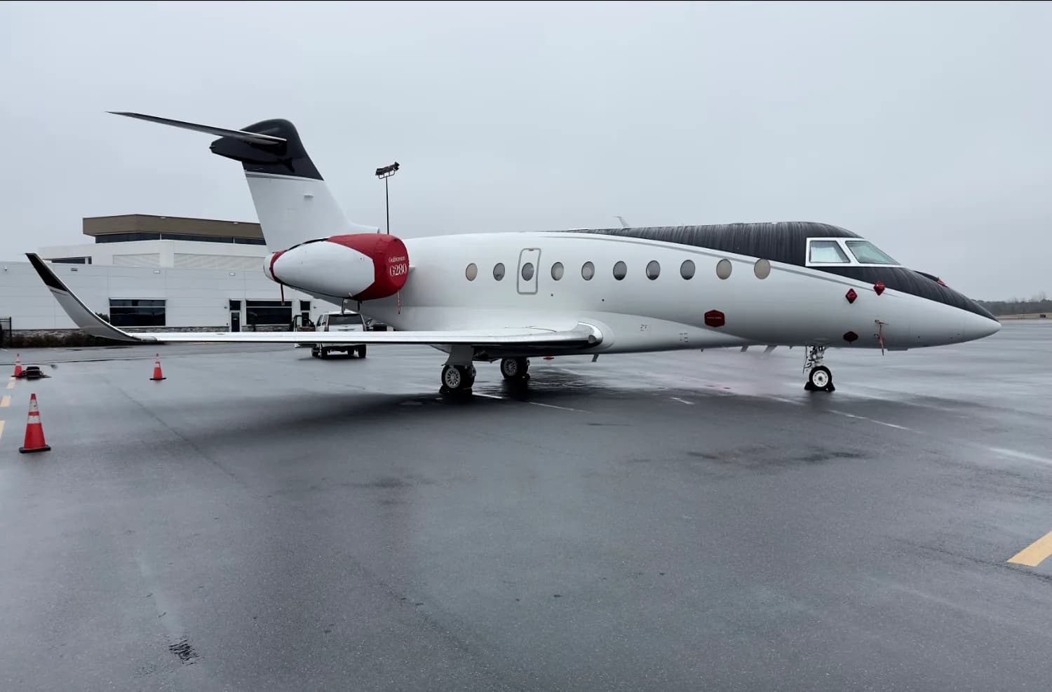 A white and black Gulfstream G280 business jet with red accents on the tail and engine nacelle, parked on a wet tarmac.