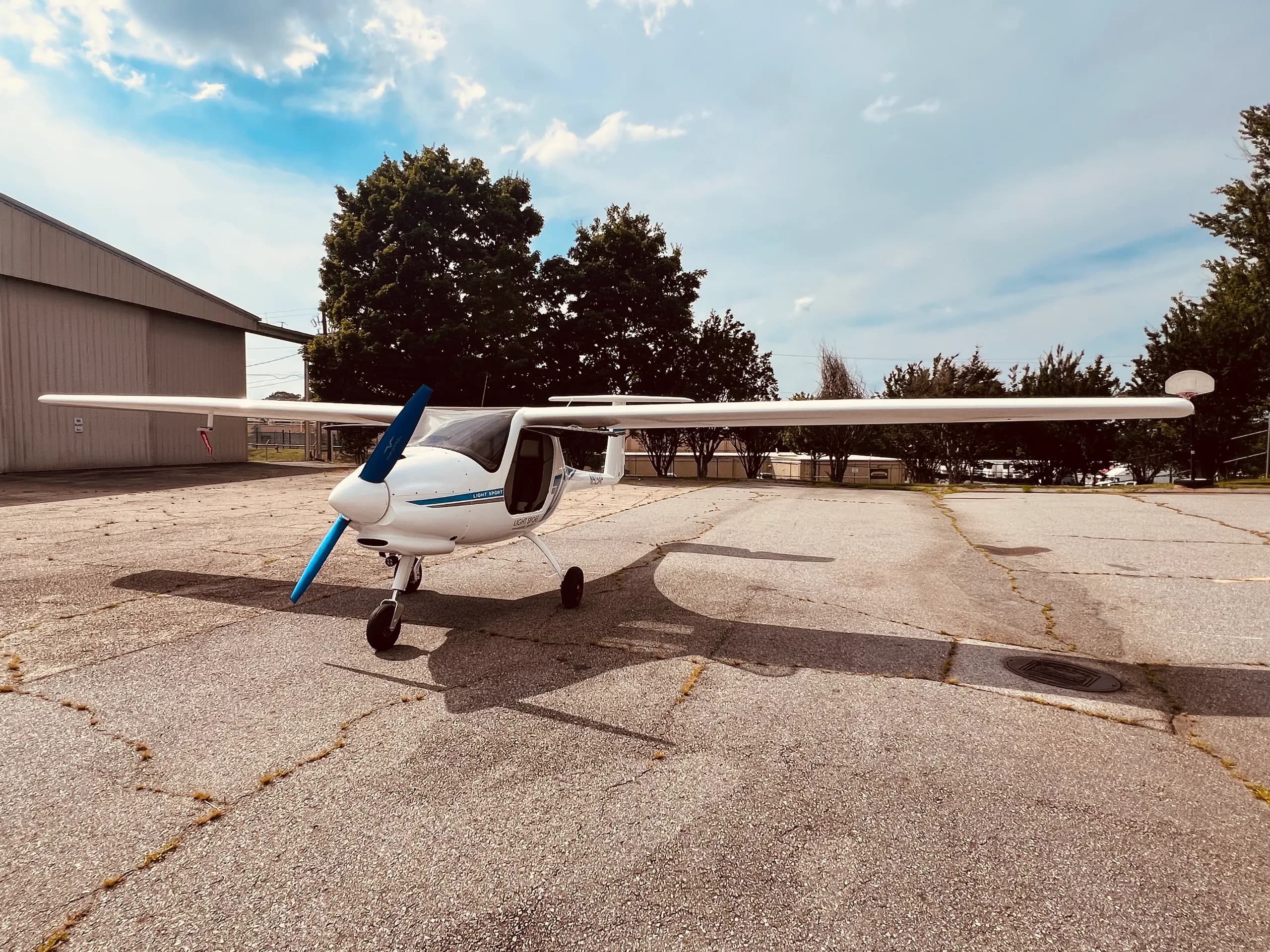 A white light sport aircraft with a blue propeller and blue stripes is parked on an asphalt apron with a hangar and trees in the background.