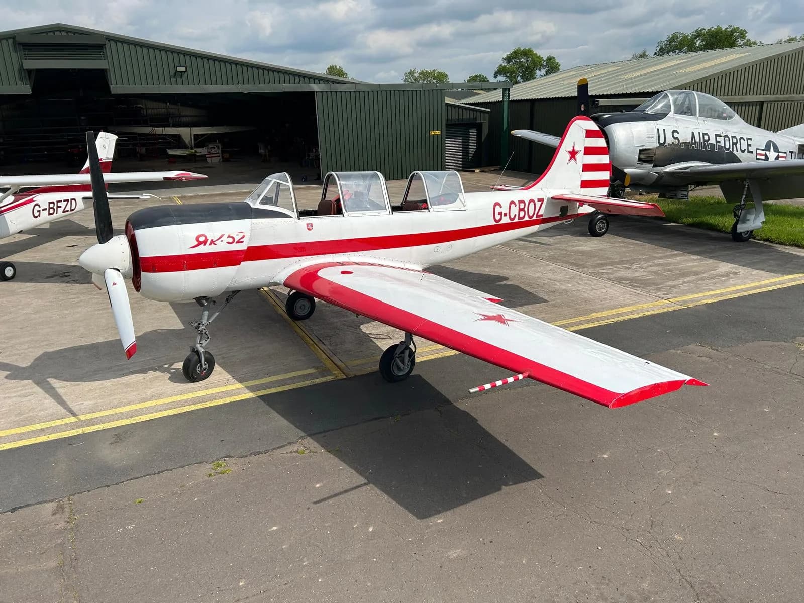 A white and red Yakovlev Yak-52 aircraft, registration G-CBOZ, is parked on an asphalt tarmac with hangars and a silver U.S. Air Force T-28 Trojan in the background.