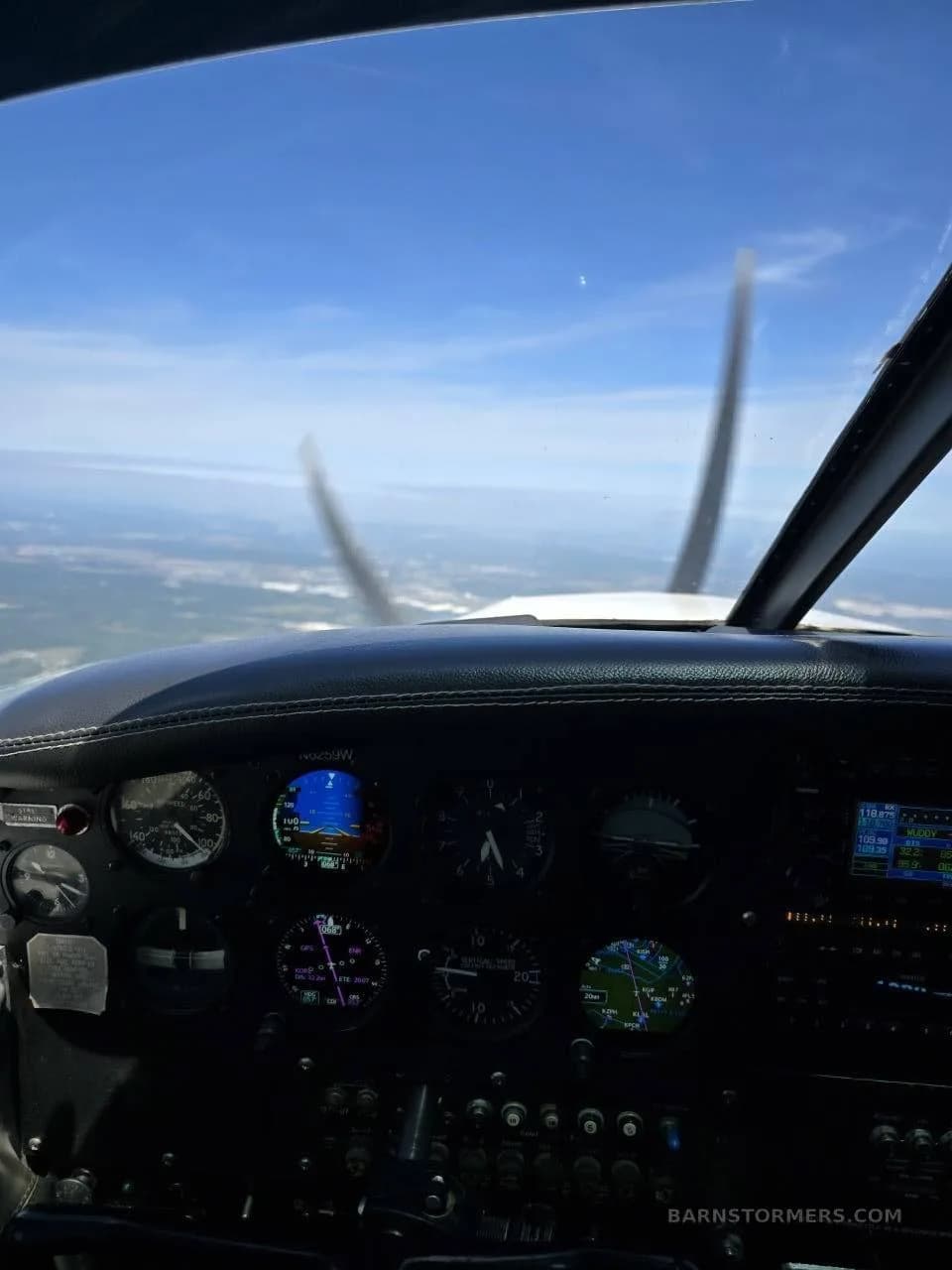 View from the cockpit of a light aircraft in flight, showing the instrument panel and propeller against a blue sky.